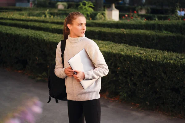 Krásná ležérní studentka v útulném svetru s notebookem a batohem zamyšleně odvracející venkovní — Stock fotografie