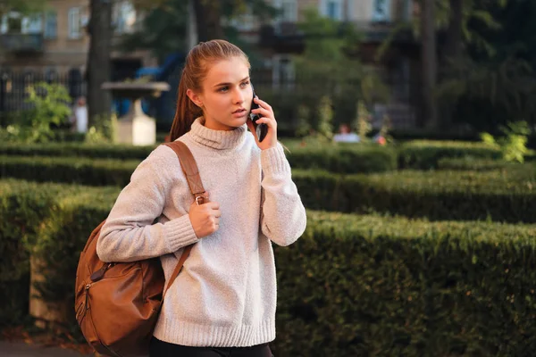 Atraktivní ležérní studentka v útulném svetru s batohem zamyšleně mluví na mobilní telefon venku — Stock fotografie