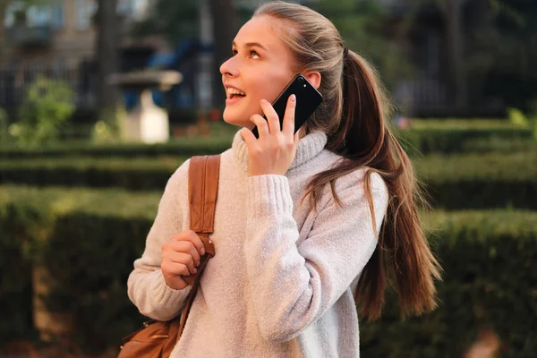 Widok z boku ładna uczennica w przytulnym swetrze z plecakiem szczęśliwie rozmawiać na zewnątrz telefon komórkowy — Zdjęcie stockowe