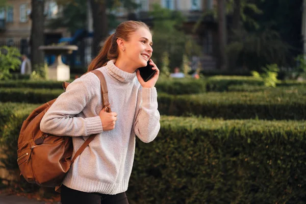 Seitenansicht des attraktiven lässigen Studentenmädchens im gemütlichen Pullover mit Rucksack, das freudig auf dem Handy im Freien spricht — Stockfoto