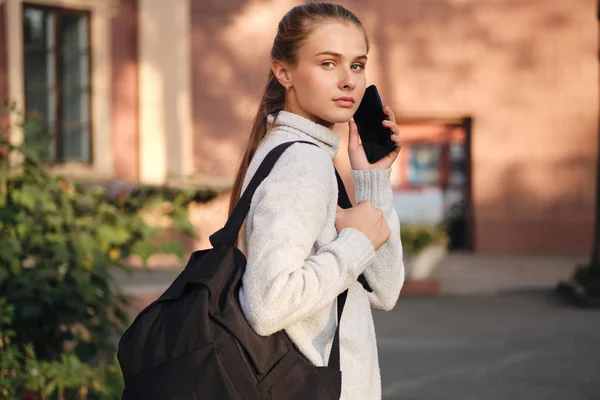 Menina estudante casual bonita em camisola aconchegante com mochila confiantemente olhando na câmera falando no celular ao ar livre — Fotografia de Stock