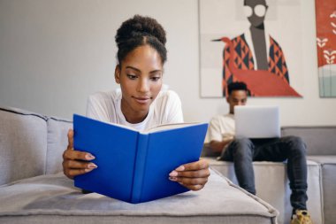 Güzel Afrikalı Amerikalı kız kanepeye uzanıp kitap okurken erkek arkadaş arka planda modern evde dizüstü bilgisayarda çalışıyor.
