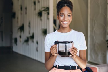 Genç, güzel, gülümseyen Afrikalı Amerikalı kadın kahve makinesiyle modern çalışma alanında kameraya neşeyle bakıyor.