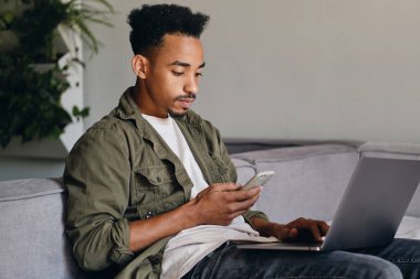 Bilgisayarlı, ciddi bir Afrikalı Amerikalı genç adam modern çalışma alanında cep telefonunu incelikle kullanıyor.