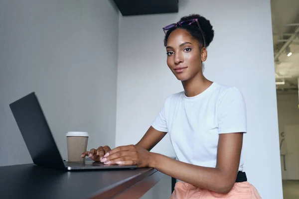 Vonzó alkalmi afro-amerikai lány dolgozik laptop kávéval menni magabiztosan keres a kamera a modern irodában — Stock Fotó