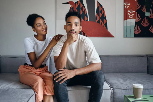 Fiatal jóképű, alkalmi afro-amerikai férfi figyelmes tekintettel a kamerába, miközben a barátnője álmodozva nézi őt a kanapén a modern otthon — Stock Fotó
