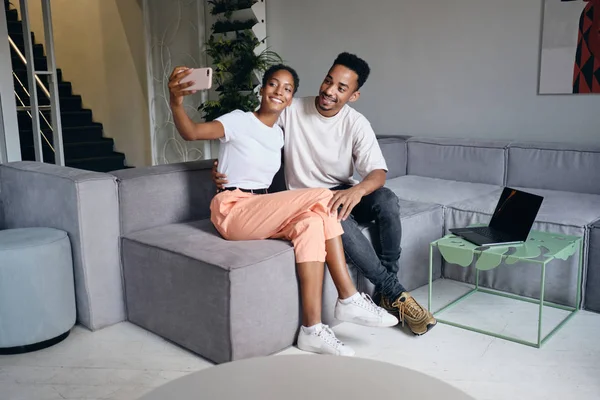 Fiatal gyönyörű alkalmi afro-amerikai pár boldogan szelfiznek mobilon együtt a kanapén a modern otthonban — Stock Fotó
