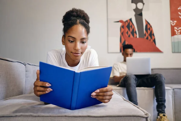 Gyönyörű afro-amerikai lány fekszik a kanapén álmodozva olvasás könyvet, míg a barátja a háttérben dolgozik laptop modern otthon — Stock Fotó