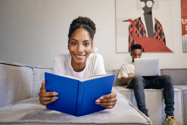 Gyönyörű mosolygós afro-amerikai lány fekszik a kanapén könyv boldogan néz a kamerába, míg a barátja a háttérben dolgozik laptop modern otthon — Stock Fotó