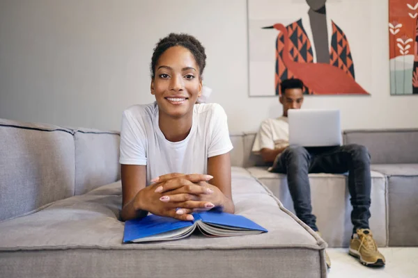 Csinos mosolygós afro-amerikai lány fekszik a kanapén könyv örömmel néz a kamerába, míg a barátja a háttérben dolgozik laptop modern otthon — Stock Fotó