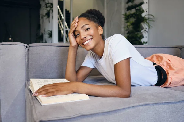 Atraktivní veselá afro americká dívka ležící na pohovce s knihou šťastně dívá do kamery v moderním domě — Stock fotografie