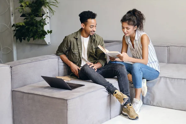 Young African American man and pretty Asian woman happily reading book together in modern co-working space — Stock Photo, Image