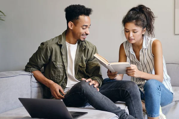 Молодой улыбающийся афроамериканец и красивая азиатка мечтательно читают вместе книгу в современном коворкинг-пространстве — стоковое фото
