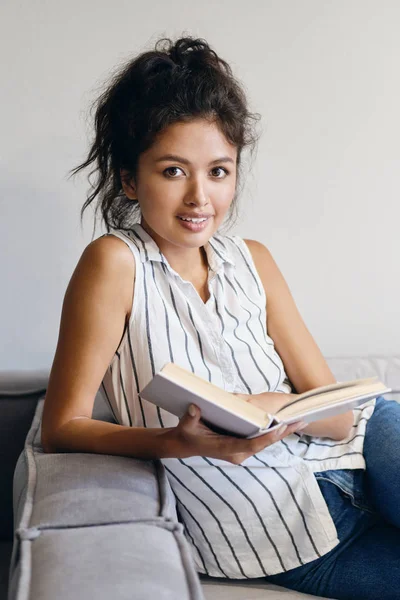 Привлекательная улыбающаяся случайная азиатская девушка с книгой, счастливо смотрящая в камеру в современном доме — стоковое фото