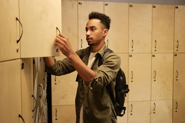 Joven hombre afroamericano casual serio con mochila dejando sus cosas en el casillero en el guardarropa de la oficina — Foto de Stock
