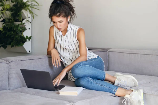 Krásná sebevědomá asijské dívka zamyšleně pracuje na notebook na pohovce v moderním domě — Stock fotografie