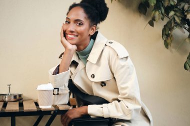 Beautiful smiling African American girl in stylish trench coat joyfully looking in camera with coffee to go in cafe on street