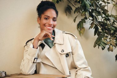 Cheerful African American girl in stylish trench coat joyfully looking in camera in cafe on street