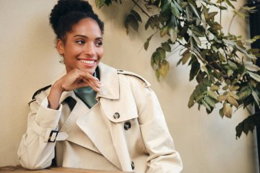 Beautiful cheerful African American girl in stylish trench coat happily looking away in cafe on street