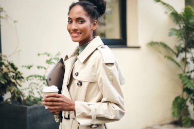 Attractive casual African American student girl in stylish trench coat with laptop and coffee to go happily looking in camera outdoor