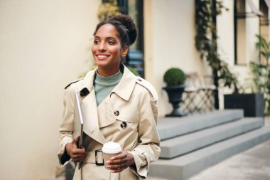 Beautiful casual African American business woman in stylish trench coat with laptop and coffee to go confidently walking down the street