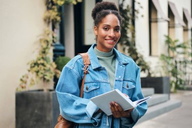 Pretty casual African American student girl in denim jacket with backpack and book happily looking in camera on city street clipart