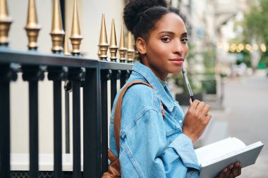 Güzel Afro-Amerikan öğrenci kız kot ceketli ve not defteriyle düşünceli bir şekilde şehir sokaklarında kameraya bakıyor.