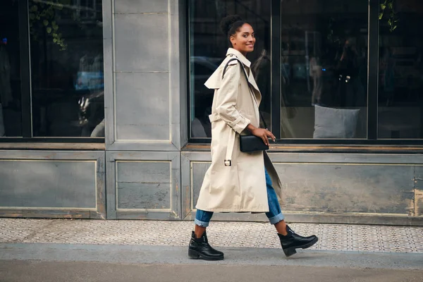 Vista lateral de sonriente chica afroamericana casual en gabardina felizmente mirando en cámara caminando por la calle — Foto de Stock