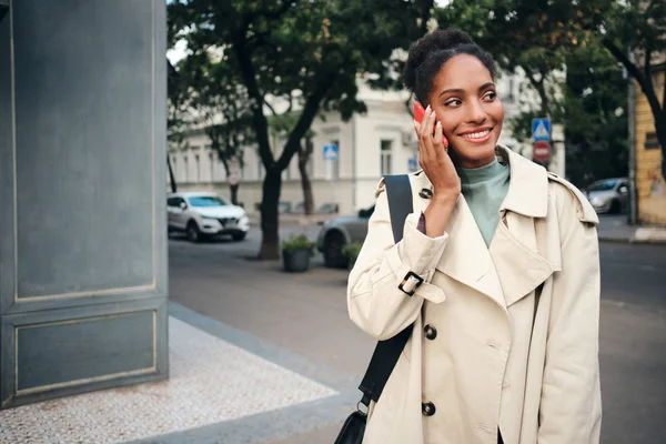 Krásná usměvavá ležérní afroamerická dívka ve stylovém plášti vesele mluvící na mobilu na ulici — Stock fotografie