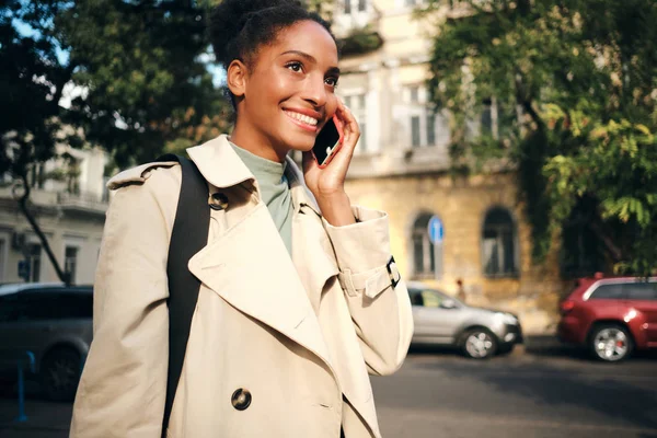Veselý Afroameričanky dívka ve stylovém trenč kabát šťastně mluví na mobilu na ulici — Stock fotografie