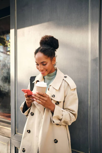 Kaunis Afrikkalainen amerikkalainen tyttö tyylikäs trenssi takki kahvia mennä iloisesti käyttäen matkapuhelin ulkona — kuvapankkivalokuva