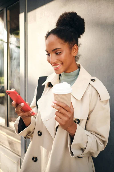 Krásná usmívající se Afroameričanka ve stylovém plášti s kávou jít šťastně pomocí mobilního telefonu venku — Stock fotografie