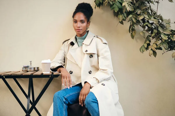 Belle fille afro-américaine décontractée en élégant trench coat regardant sensuellement à la caméra dans un café dans la rue — Photo