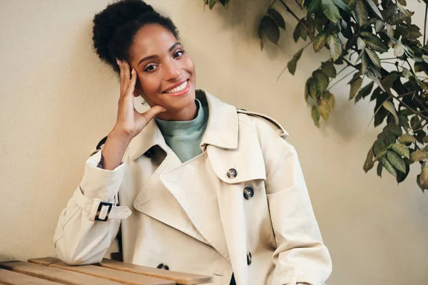 Pretty smiling casual African American girl in stylish trench coat joyfully looking in camera in cafe on street — 图库照片