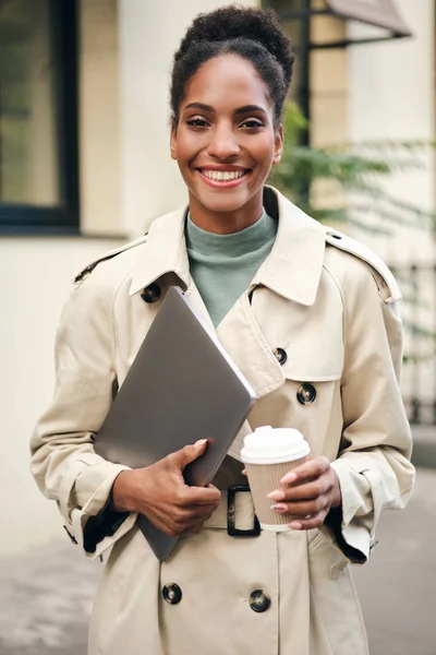 Cheerful casual African American business woman in stylish trench coat with laptop and coffee to go joyfully looking in camera outdoor — ストック写真