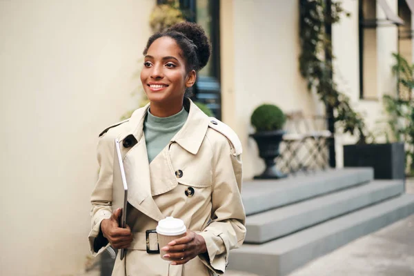 Beautiful casual African American business woman in stylish trench coat with laptop and coffee to go confidently walking down the street — Stock fotografie