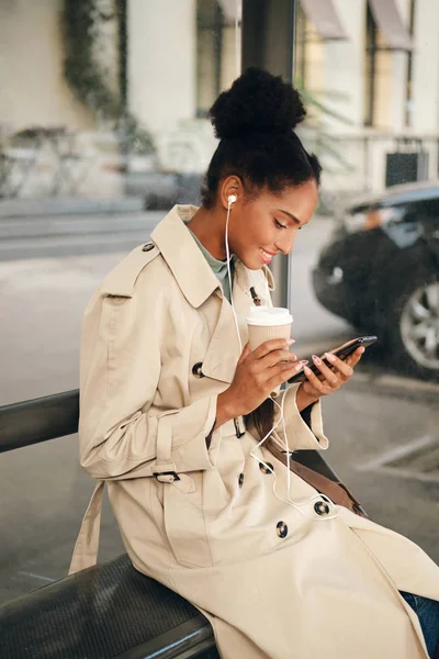 Side näkymä hymyilevä rento Afrikkalainen Amerikan tyttö tyylikäs trenssi takki ja kuulokkeet kahvia mennä iloisesti käyttäen matkapuhelin bussipysäkillä — kuvapankkivalokuva