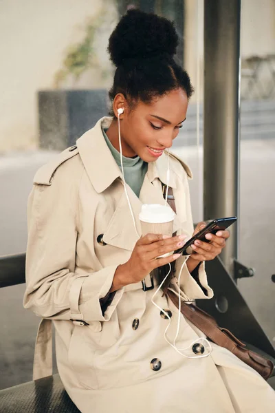 멋진 해구 코트와 커피가든 이어폰을 입고 버스 정류장에서 핸드폰을 행복하게 사용하는 평범 한 아프리카 계 미국인 소녀 — 스톡 사진