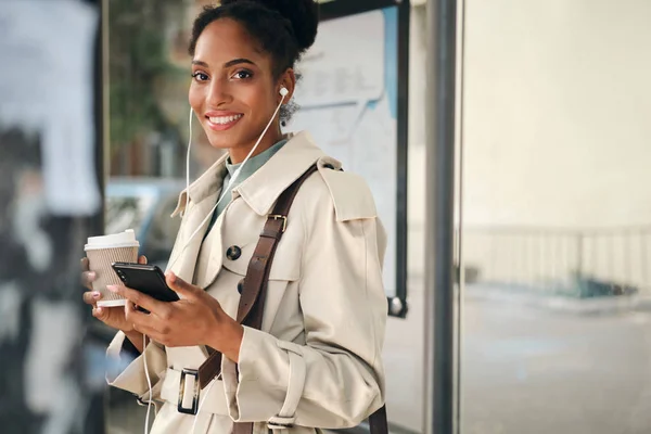 Krásná ležérní afro americká dívka ve stylovém plášti s kávou jít a mobilní telefon radostně dívá do kamery na autobusové zastávce — Stock fotografie