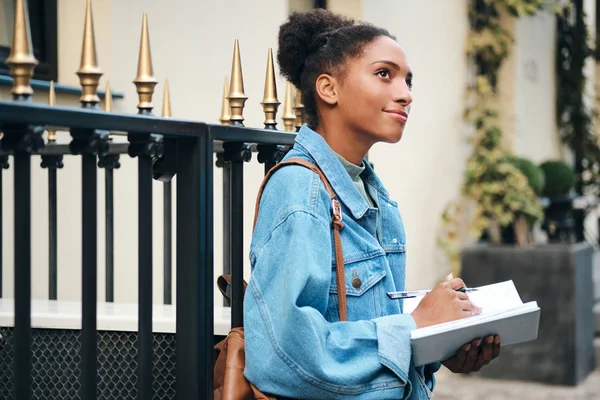 Side view of beautiful casual African American student girl w dżinsowej kurtce z plecakiem drethe writing notes on city street — Zdjęcie stockowe