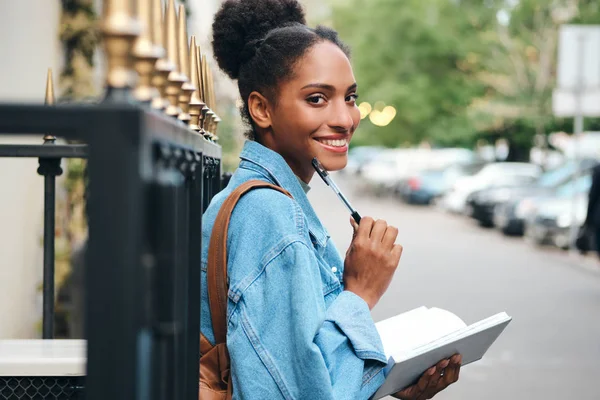 Vidám alkalmi afro-amerikai diáklány farmerdzsekiben, jegyzetfüzettel, boldogan néz a kamerába a város utcáján. — Stock Fotó