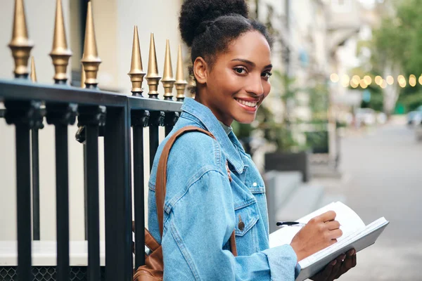 Gyönyörű alkalmi afro-amerikai diák lány farmer kabát jegyzettömbbel örömmel nézett a kamera a város utcájában — Stock Fotó