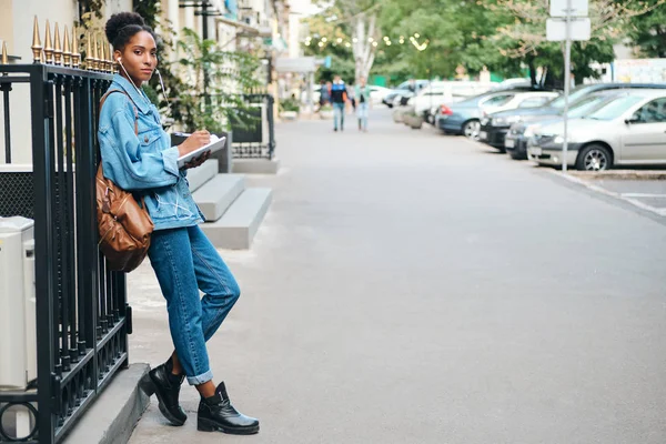 Vista lateral da menina estudante afro-americana casual em jaqueta jeans com mochila e bloco de notas olhando com confiança na câmera na rua da cidade — Fotografia de Stock