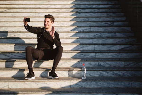 Pemuda tampan tersenyum sporty pria gembira menunjukkan sikap hai berbicara di video chat di ponsel setelah olahraga di luar ruangan — Stok Foto