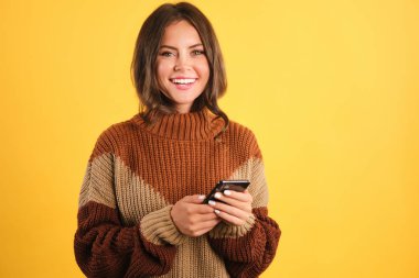 Hoş süveter giyen, cep telefonlu, sarı arka planda kameraya bakan çekici, neşeli bir kız.