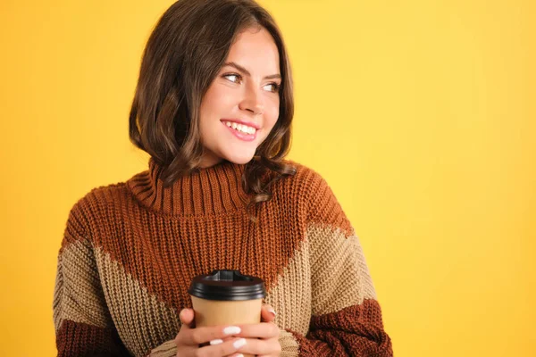 Beautiful smiling girl in cozy sweater with cup to go joyfully looking aside over yellow background — Stock Photo, Image