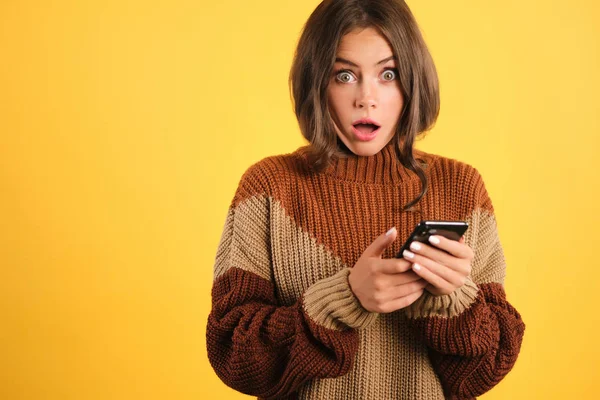 Chica aturdida en suéter de punto con el teléfono celular asombradamente mirando en la cámara sobre fondo amarillo — Foto de Stock