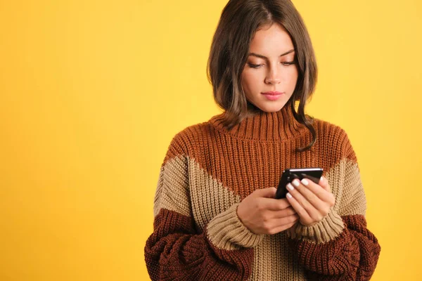 Kaunis tyttö viihtyisä villapaita luottavaisin mielin käyttämällä matkapuhelinta keltaisella taustalla — kuvapankkivalokuva