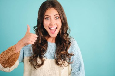 Oldukça heyecanlı bir kız, hoş bir süveter içinde, mavi arka planda başparmak hareketi yapıyor.