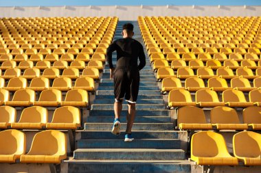 Genç ve güçlü Afrikalı Amerikalı sporcunun boş stadyum koltukları arasında koşuşunun arka görüntüsü 